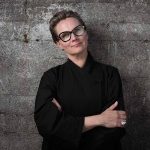 Skilled female chef with glasses, confident pose, dark kitchen uniform, inspiring culinary professional.