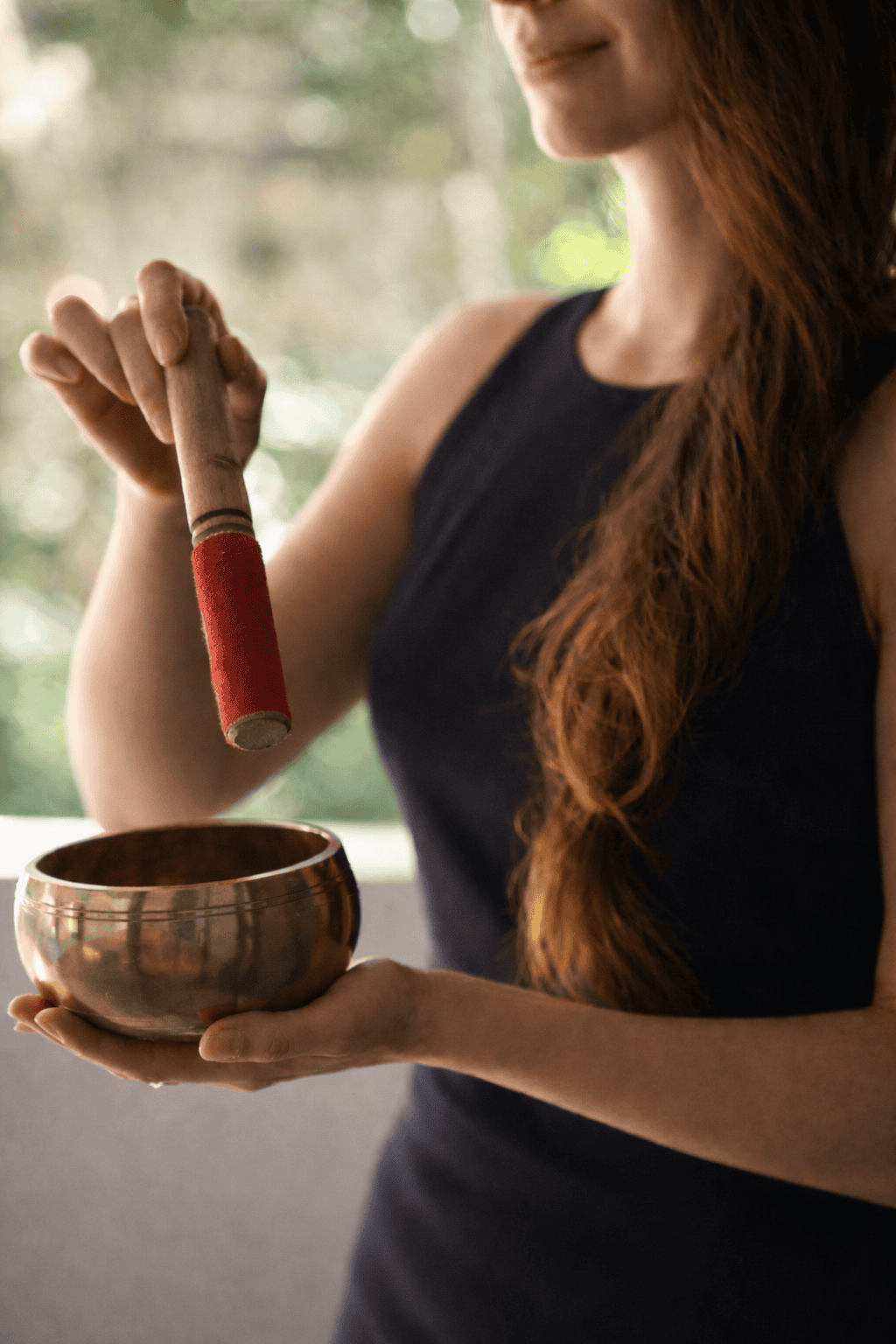 Relaxed woman holding a singing bowl and a large wooden mallet, practicing sound healing meditation.