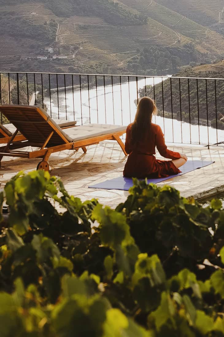 Tranquil woman meditating on balcony overlooking river and vineyards in scenic countryside.
