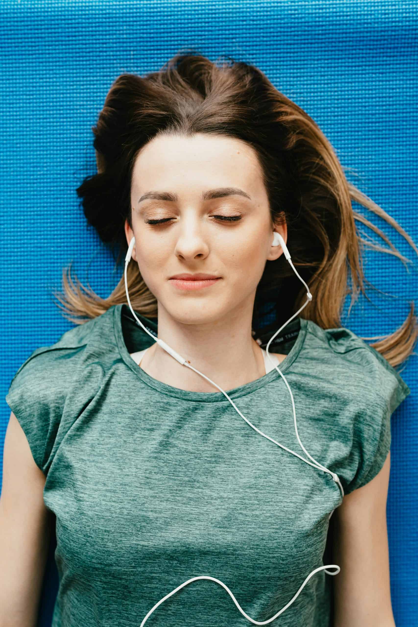 Relaxed young woman listening to music with earbuds, lying on a blue mat.